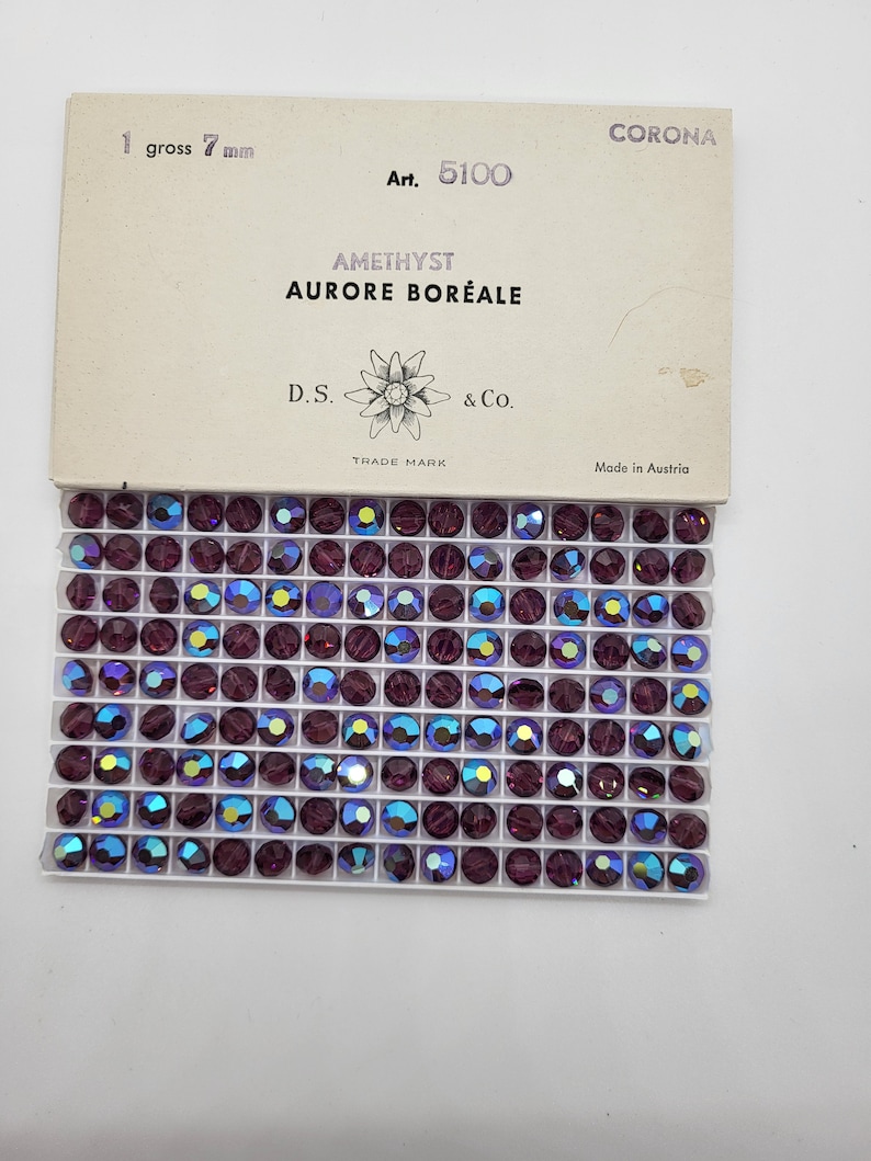 May include: A box of amethyst-coloured, iridescent rhinestones, each 7 mm in diameter, arranged in a grid. The package includes text that reads "AMETHYST AURORE BOR&Eacute;ALE" and "Made in Austria."