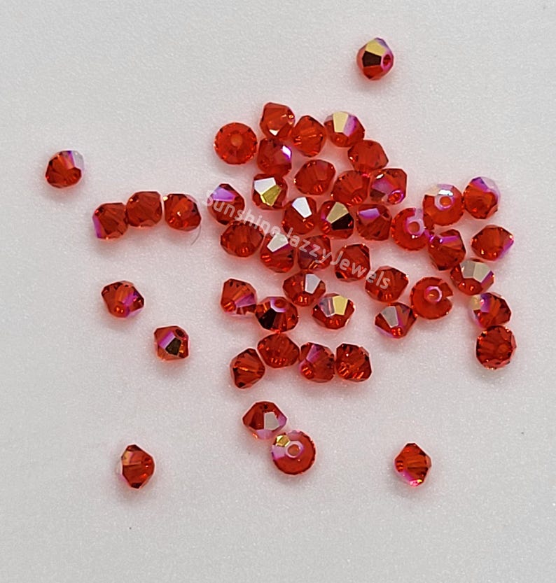 K&ouml;nnte beinhalten: Eine Ansammlung kleiner, facettierter, bikonischer Perlen in einem leuchtenden Orange-Rot. Jede Perle hat ein mittiges Loch und eine irisierende Oberfl&auml;che, die das Licht einf&auml;ngt und subtile Farbver&auml;nderungen auf den Glasperlen erzeugt.