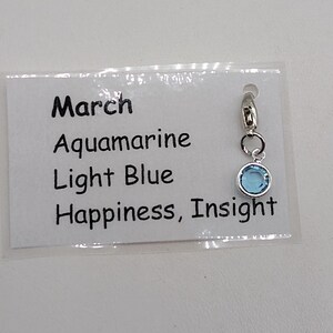 May include: A light blue charm with a silver clasp. The charm is labeled "March", "Aquamarine", "Light Blue", and "Happiness, Insight".