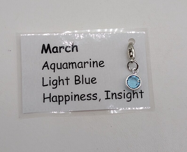 May include: A light blue charm with a silver clasp. The charm is labeled "March", "Aquamarine", "Light Blue", and "Happiness, Insight".