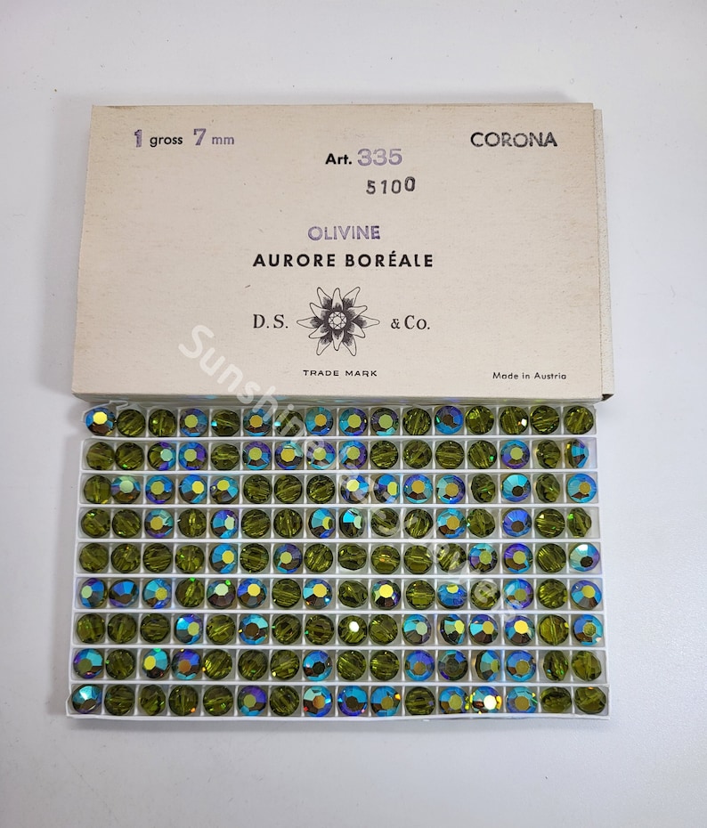 K&ouml;nnte beinhalten: Eine Schachtel mit 7 mm gro&szlig;en gr&uuml;nen Glasperlen mit einem Etikett mit der Aufschrift "CORONA Art. 335 5100 OLIVINE AURORE BOR&Eacute;ALE D.S. & Co. TRADE MARK Made in Austria".