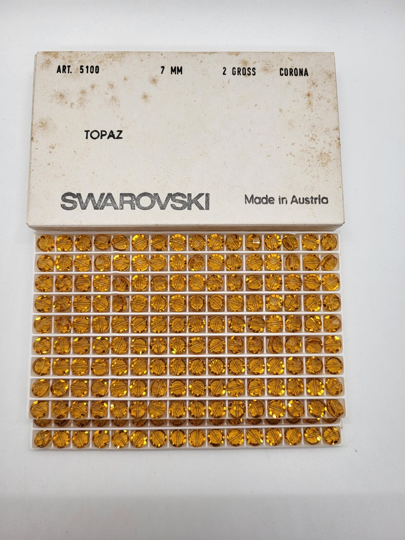 Puede incluir: Primer plano de una caja de cristales Swarovski topacio de 7 mm. La caja est&aacute; etiquetada con "ART. 5100", "7 MM", "2 GROSS", "CORONA", "TOPAZ" y "SWAROVSKI". La caja tambi&eacute;n indica "Made in Austria".
