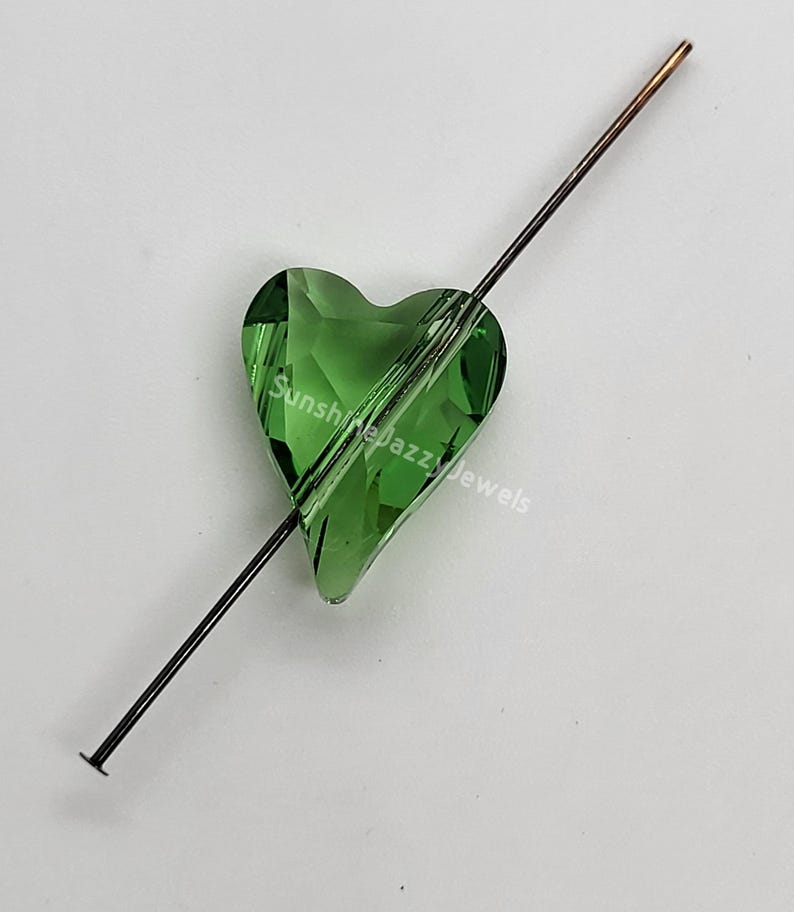 May include: A green crystal heart bead with a hole for stringing. The bead is on a thin metal wire.