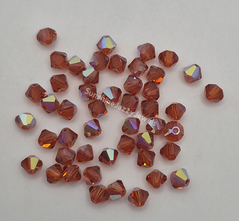 May include: A scattering of faceted, reddish-brown beads with iridescent highlights. The beads are small, with multiple facets that catch the light, creating a shimmering effect. The beads are likely for jewelry making or crafts.