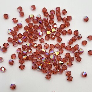 May include: A scattering of faceted, reddish-brown beads with iridescent highlights. The beads are small, square-cut, and reflect light with a rainbow sheen. They are scattered across a white surface, suggesting a craft or jewelry-making theme.