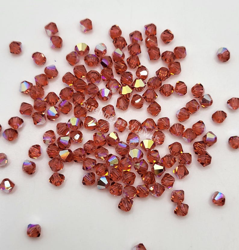May include: A scattering of faceted, reddish-brown beads with iridescent highlights. The beads are small, square-cut, and reflect light with a rainbow sheen. They are scattered across a white surface, suggesting a craft or jewelry-making theme.