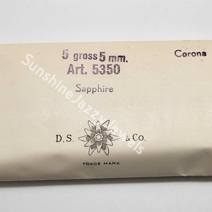 May include: A white paper package with the text "5 gross 5 mm. Art. 5350 Corona Sapphire D.S. & Co. TRADE MARK" printed on it. The package features a stylized flower with a star in the center.