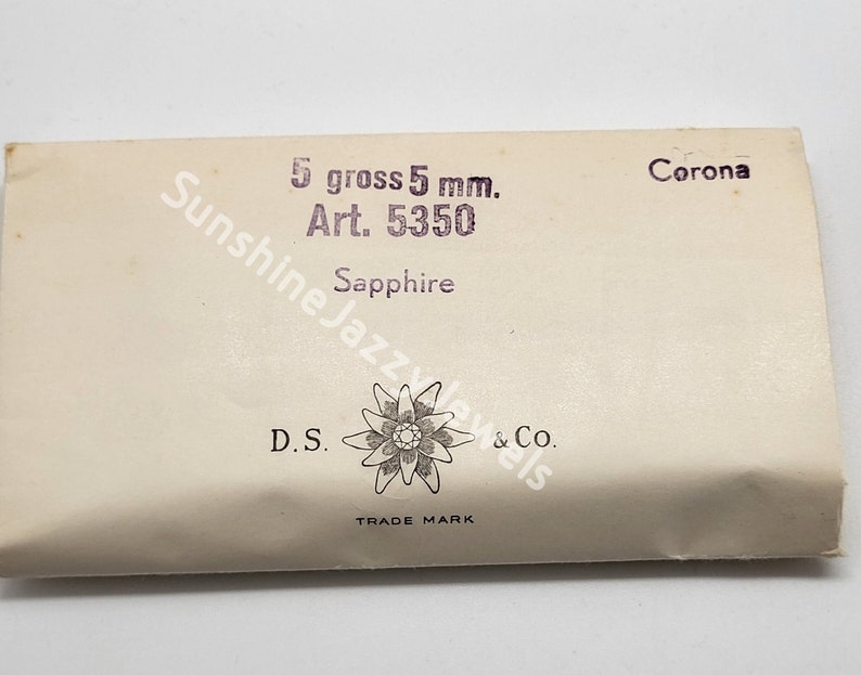 May include: A white paper package with the text "5 gross 5 mm. Art. 5350 Corona Sapphire D.S. & Co. TRADE MARK" printed on it. The package features a stylized flower with a star in the center.