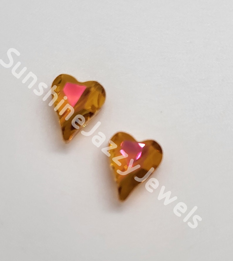 K&ouml;nnte beinhalten: Zwei goldene herzf&ouml;rmige Strasssteine mit einem pinkfarbenen, schillernden Zentrum.