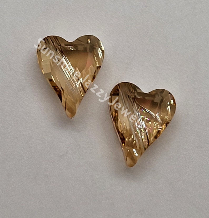 K&ouml;nnte beinhalten: Zwei goldene herzf&ouml;rmige Kristallperlen mit einem gestreiften Muster. Die Perlen befinden sich auf einem wei&szlig;en Hintergrund.