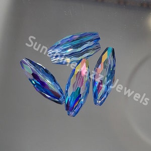 May include: Four faceted, elongated crystal beads in shades of blue, with iridescent and rainbow reflections. The beads are arranged on a reflective surface. The text "Sunshine Dazzy Jewels" is visible in the image.