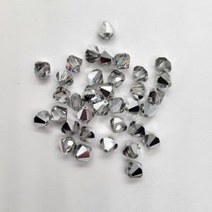 May include: A group of silver-toned faceted glass beads, also known as bicones, scattered on a white surface. The beads have a pointed shape and are designed for jewelry making.