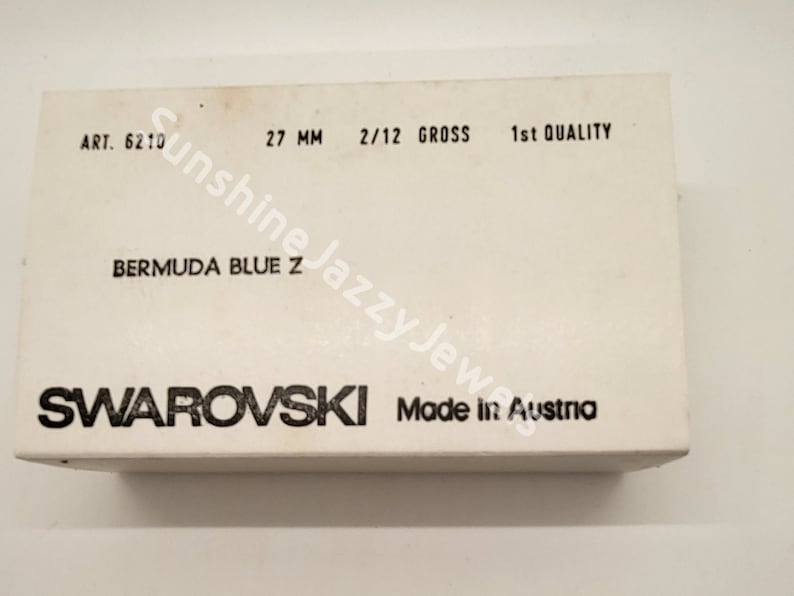May include: A white box with black text that reads "ART. 6210 27 MM 2/12 GROSS 1st QUALITY BERMUDA BLUE Z SWAROVSKI Made in Austria".
