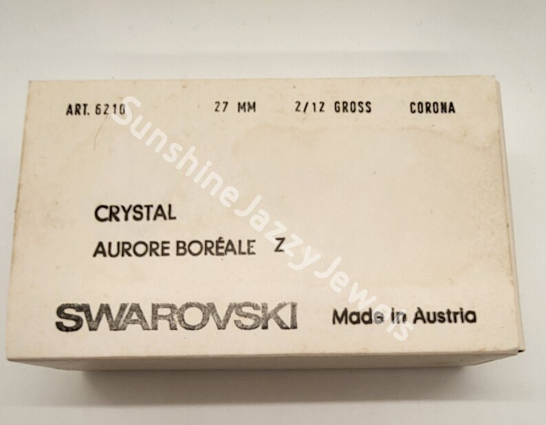 May include: A white box with black text that reads "SWAROVSKI Made in Austria" and "CRYSTAL AURORE BOR&Eacute;ALE". The box also has the text "ART. 6210", "27 MM", "2/12 GROSS", and "CORONA".