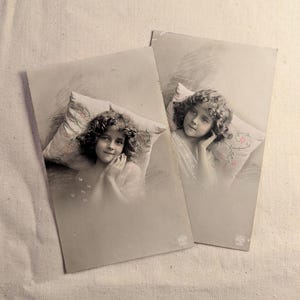 Puede incluir: Dos postales vintage en tonos sepia con retratos de una niña con cabello rizado. Descansa sobre una almohada con bordados florales. Las postales están ligeramente anguladas, mostrando la textura del papel envejecido y una sutil marca de agua.