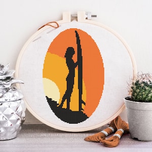 May include: A cross-stitch embroidery hoop with a sunset scene featuring a silhouette of a person standing with a surfboard. The background is a gradient of orange, yellow, and white.