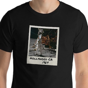 May include: A black t-shirt with a vintage-style photo of an astronaut on the moon. The photo has the text "HOLLYWOOD CA 1969" written on it.