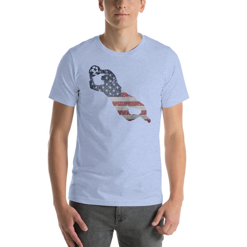 Puede incluir: Una camiseta azul claro con la silueta de un jugador de f&uacute;tbol atrapando un bal&oacute;n. La silueta est&aacute; llena con un dise&ntilde;o de bandera estadounidense.