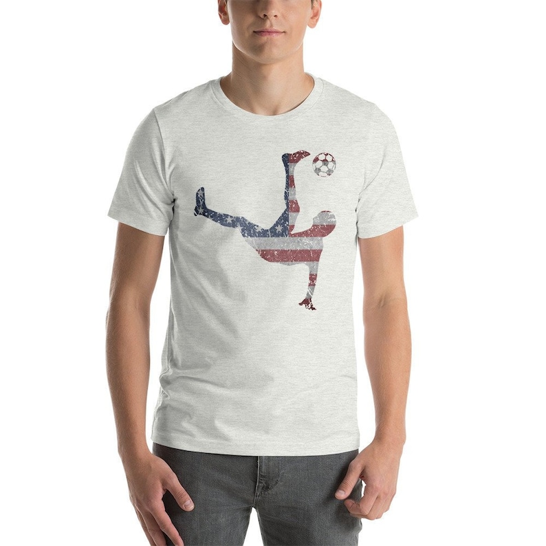K&ouml;nnte beinhalten: Ein hellgraues T-Shirt mit der Silhouette eines Fu&szlig;ballspielers, der einen Ball tritt. Der Spieler ist auf ein amerikanisches Flaggen-Design gelegt.