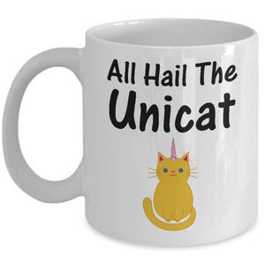 Puede incluir: Taza de café de cerámica blanca con una ilustración en blanco y negro de un gato con un cuerno de unicornio. El texto "All Hail The Uniccat" está impreso en la taza.