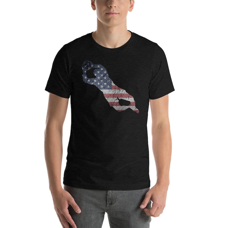 Puede incluir: Una camiseta negra con un dise&ntilde;o de bandera estadounidense desgastado en forma de mano haciendo el signo 'OK'.