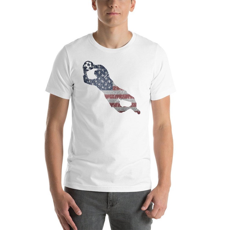 Puede incluir: Camiseta blanca con la silueta de un portero de f&uacute;tbol atrapando un bal&oacute;n. El portero est&aacute; delineado con la bandera estadounidense.