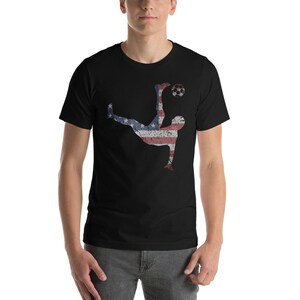K&ouml;nnte beinhalten: Ein schwarzes T-Shirt mit einer Silhouette eines Fu&szlig;ballspielers, der einen Ball tritt. Der Spieler ist in den Farben der amerikanischen Flagge umrandet.