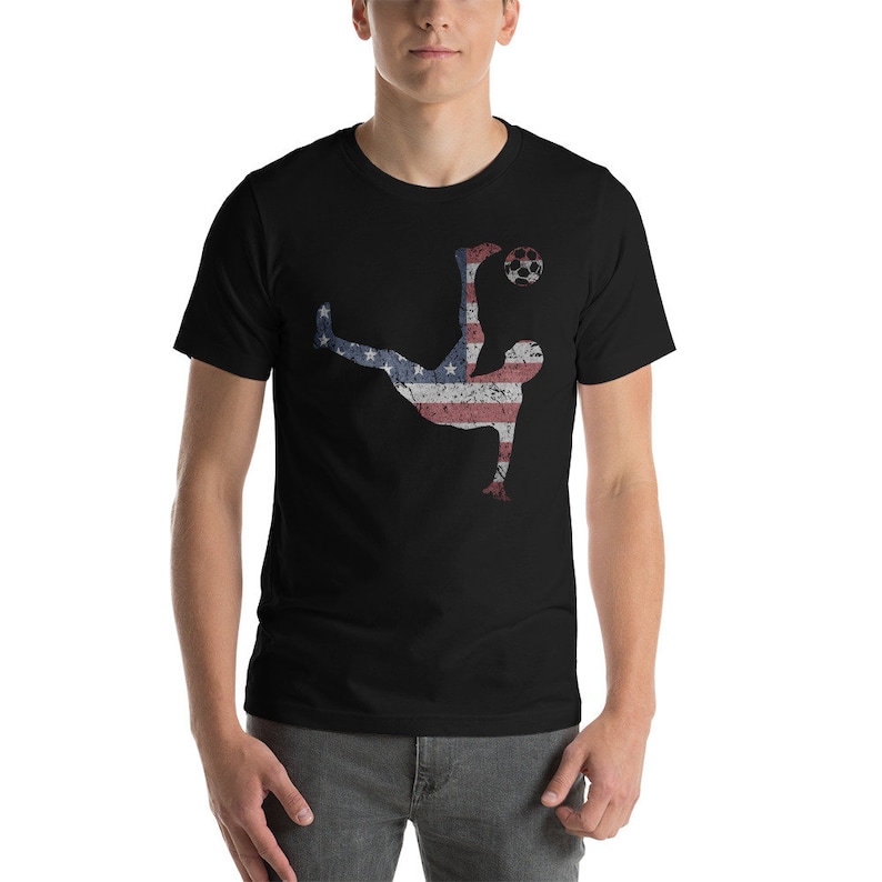 K&ouml;nnte beinhalten: Ein schwarzes T-Shirt mit einer Silhouette eines Fu&szlig;ballspielers, der einen Ball tritt. Der Spieler ist in den Farben der amerikanischen Flagge umrandet.