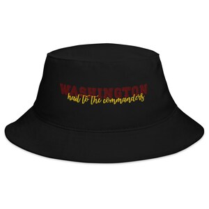 Op de afbeelding: Een zwarte bucket hat met geborduurde tekst op de voorzijde. Het woord WASHINGTON is gestikt in donkerrode hoofdletters, met de zin hail to the commanders er direct onder geborduurd in een geel cursief lettertype.