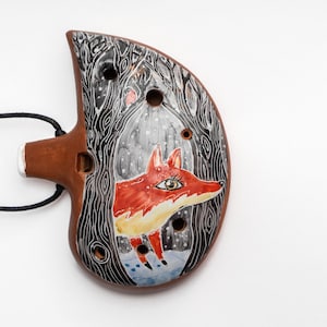 Peut inclure: Une ocarina en céramique avec une scène de forêt noire et blanche et un renard rouge peint dessus. L'ocarina a un cordon noir pour la porter autour du cou.