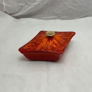 May include: A red ceramic dish with a lid. The lid has an orange and yellow floral design and a gold-coloured knob. The dish is rectangular with rounded corners. The lid's design resembles a flower.