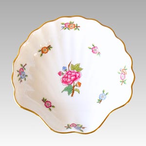 May include: A white, shell-shaped porcelain dish with a scalloped gold rim. The dish is decorated with colorful floral patterns, including a large pink peony in the center and smaller flowers in shades of pink, blue, and orange.