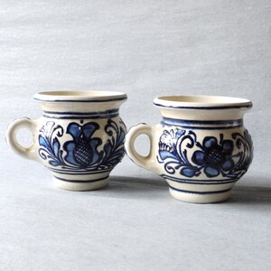 May include: Two ceramic mugs with a cream-colored base and blue floral designs. Each mug has a small handle and a blue rim. The floral pattern includes stylized flowers and leaves. The mugs are displayed on a light gray surface.
