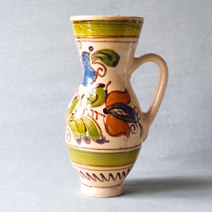 May include: A hand-painted ceramic pitcher with a handle. The pitcher features a beige base with colorful floral designs in blue, orange, and green. Green and yellow bands encircle the top and base, with a zig-zag pattern near the bottom.