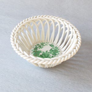 May include: A white ceramic basket with a woven design and a decorative rope-like edge. The interior base features a floral pattern in shades of green. This decorative basket is ideal for displaying small items.