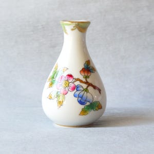 May include: A small, off-white ceramic vase with a floral design. The vase features a slender neck and a rounded body. The floral design includes pink, blue, and orange flowers with green leaves on a brown branch. The rim is trimmed with gold.
