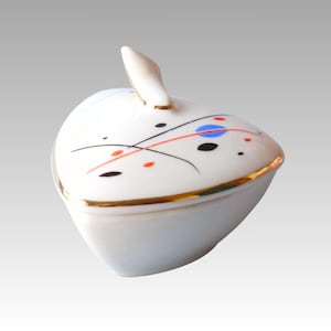 May include: A white ceramic apple-shaped trinket box with a lid. The lid is decorated with a gold rim and abstract black, red, and blue designs. A small, white, leaf-shaped handle is on top.
