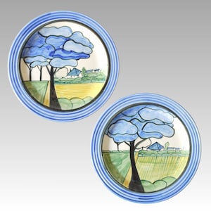 Può includere: Due piatti in ceramica blu e bianca con un design paesaggistico stilizzato. Ogni piatto presenta un albero blu con un campo giallo e una casa bianca sullo sfondo.