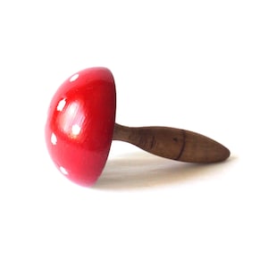 Może przedstawiać: Drewniany przedmiot w kształcie grzyba z czerwonym kapeluszem i białymi kropkami. Trzon jest brązowy. Grzyb leży na jasnej powierzchni.