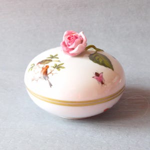 Peut inclure: Boîte à bijoux en porcelaine blanche ornée d'une rose rose sur le dessus. La boîte est décorée d'oiseaux peints et d'éléments floraux. Une bande dorée entoure la base. Le couvercle est orné d'une rose rose et de feuilles vertes.