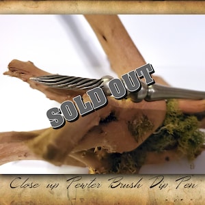 Puede incluir: Primer plano de una pluma de inmersi&oacute;n de pincel de plata que descansa sobre un trozo de madera a la deriva con musgo verde. La pluma tiene una punta de metal retorcida y un mango de plata. La imagen tiene el texto "SOLD OUT" sobre la pluma.