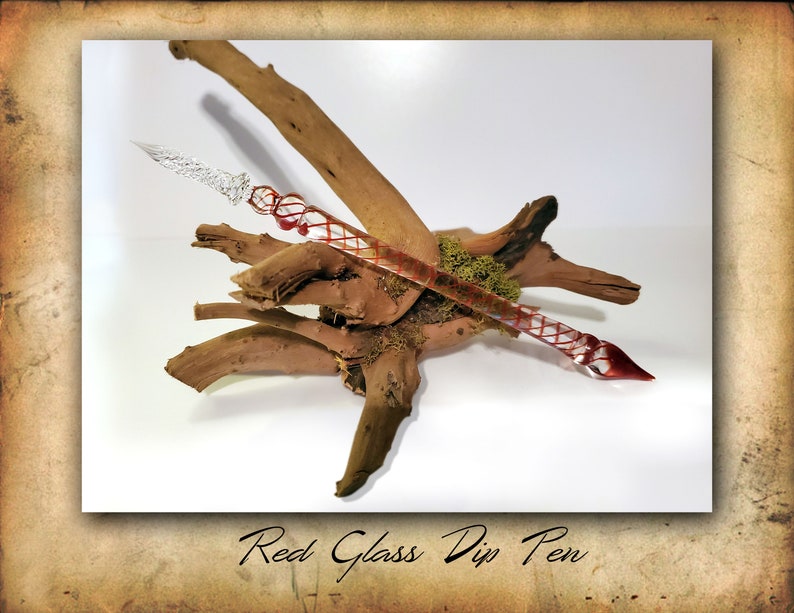 Puede incluir: Un plum&iacute;n de tinta de vidrio rojo y transparente con un dise&ntilde;o de espiral decorativo. La pluma est&aacute; apoyada en un trozo de madera a la deriva.