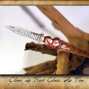 Puede incluir: Primer plano de una pluma de tinta de vidrio rojo y transparente. La pluma tiene una punta de vidrio transparente y un cuerpo de vidrio rojo con remolinos rojos y blancos.