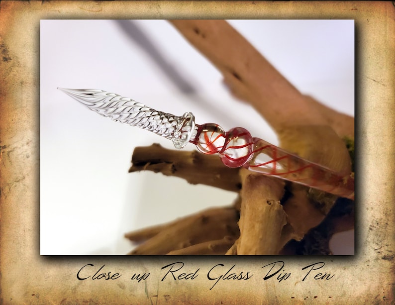 Puede incluir: Primer plano de una pluma de tinta de vidrio rojo y transparente. La pluma tiene una punta de vidrio transparente y un cuerpo de vidrio rojo con remolinos rojos y blancos.