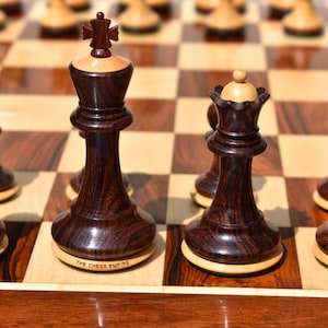 Könnte beinhalten: Nahaufnahme eines Schachbretts mit dunklen Holzfiguren. Die Figuren sind in Spielstellung angeordnet, mit dem schwarzen König und der schwarzen Dame im Vordergrund. Das Schachbrett besteht aus hellen und dunklen Holzfeldern. Der Text "The Chess Empire" ist auf der Basis des schwarzen Königs sichtbar.