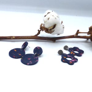 Op de afbeelding: Twee paar handgemaakte oorbellen. Eén paar is marineblauw met een cirkelvormig ontwerp en kleurrijke spikkels. Het andere paar is marineblauw met een bloemachtige vorm en kleurrijke spikkels.