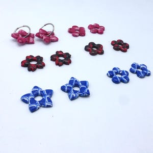 Può includere: Una collezione di componenti di gioielli a forma di fiore in vari colori. Include fiori rosa, rossi e blu, alcuni con cerchietti argentati. I fiori hanno un aspetto marmorizzato o fantasia, adatti per l'artigianato o la creazione di gioielli. Misure in centimetri.