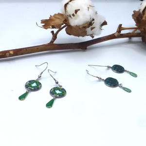 Può includere: Orecchini color argento con un design circolare verde e bianco e un pendente verde a forma di goccia. Gli orecchini sono esposti su una superficie bianca con un ramo di cotone sullo sfondo. Un accessorio unico.
