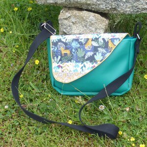 Peut inclure: Un sac à bandoulière vert turquoise avec une bandoulière noire réglable. Le rabat avant du sac est en tissu à motifs colorés sur le thème de la jungle, avec des animaux, des plantes et des feuilles.