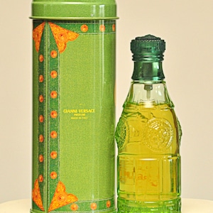 May include: A green perfume bottle and matching cylindrical container. The bottle is clear with a green cap and base, filled with yellow liquid. The container has a denim-like texture with orange and gold accents. The text "GIANNI VERSACE PROFUMI MADE IN ITALY" is visible.